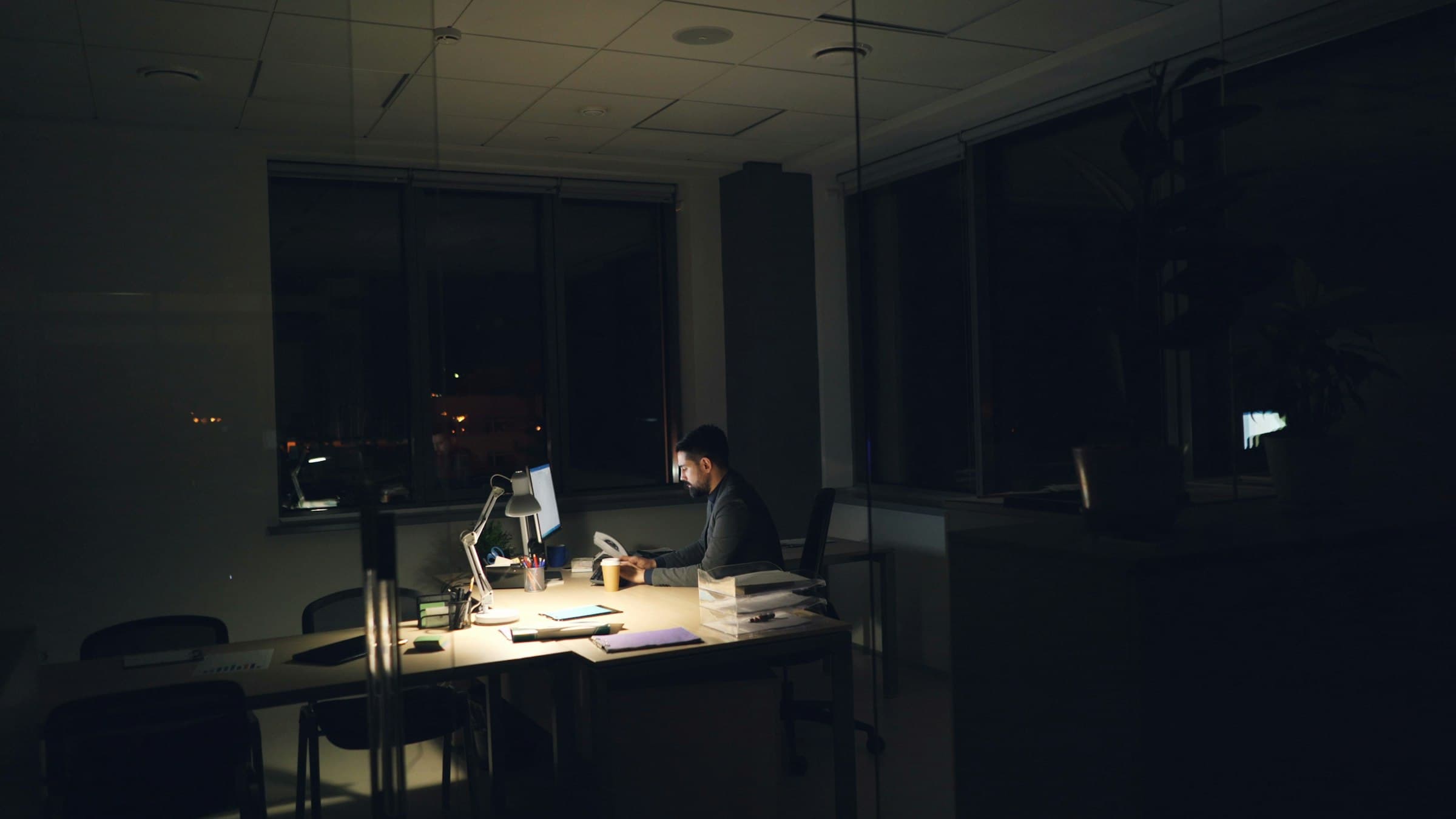 A man working late at a single lit desk in a dark office